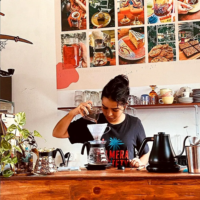 A barista in a 'Palmera Society' T-shirt pours a V60 pour-over at the café's wooden bar, under a wall of food photographs.
