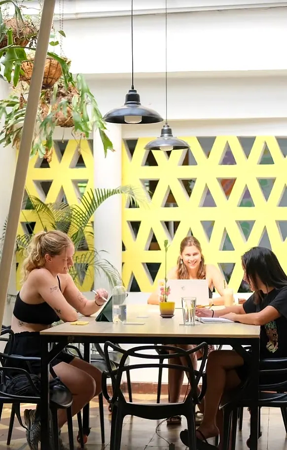 Three guests working at a shared table under pendant lamps, in front of the yellow triangular breeze-block wall.