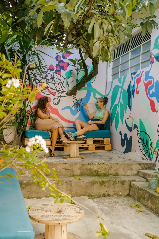 Two guests chatting on a pallet sofa in the mural-covered patio, under a tree.