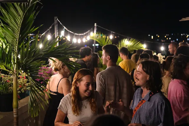 A packed rooftop night: guests talking and dancing under string lights, with city lights behind.