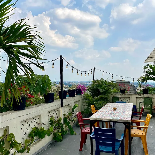 La Palmera's rooftop terrace: a mosaic-tiled communal table with red, yellow, blue and green chairs, potted palms, bougainvillea and string lights, with Cali's skyline beyond.