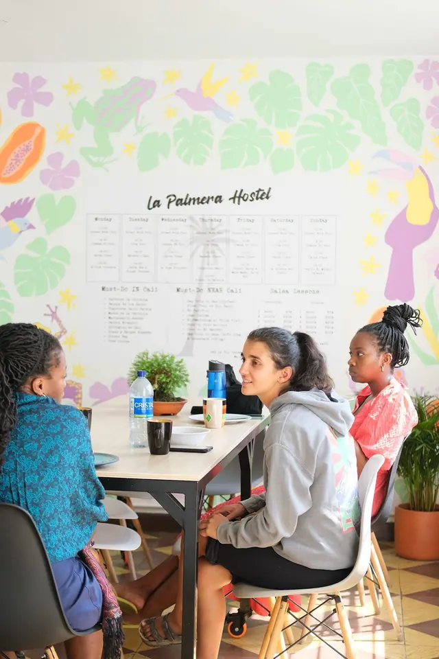 Guests talking at the dining tables in front of the hand-painted weekly activity wall, framed by monstera leaves, a toucan and a frog.