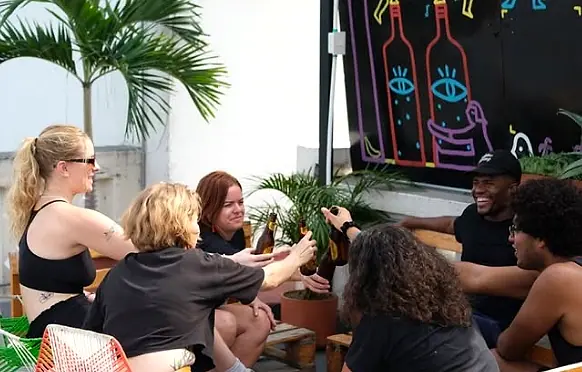 Guests sharing beers around a table on the terrace, under palms and a neon-style bottle mural.